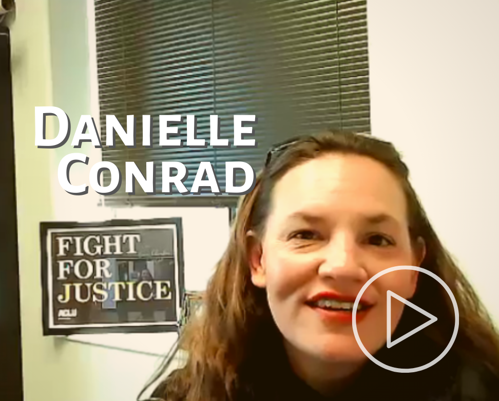 Link to Danielle Conrad's interview. She smiles into the camera with a "Fight for Justice" sign in the background.